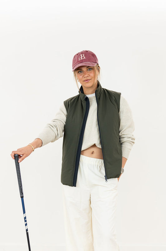 Person wearing a green vest, white shirt, and white pants with a maroon cap, holding a golf club on a white background.
