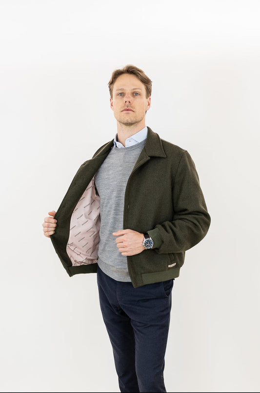 Man wearing a green khaki bomber jacket, gray sweater, dark pants, and brown shoes on a white background